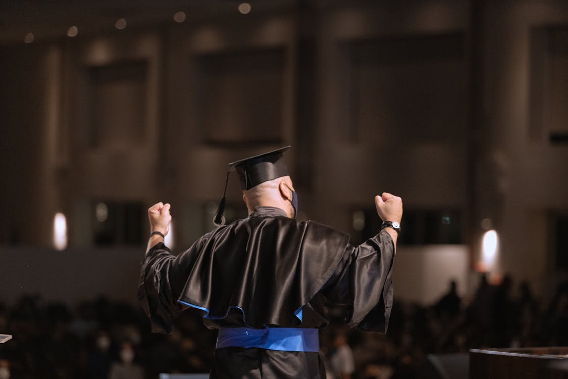 Free Graduate celebrating success at ceremony with arms raised in joy. Stock Photo