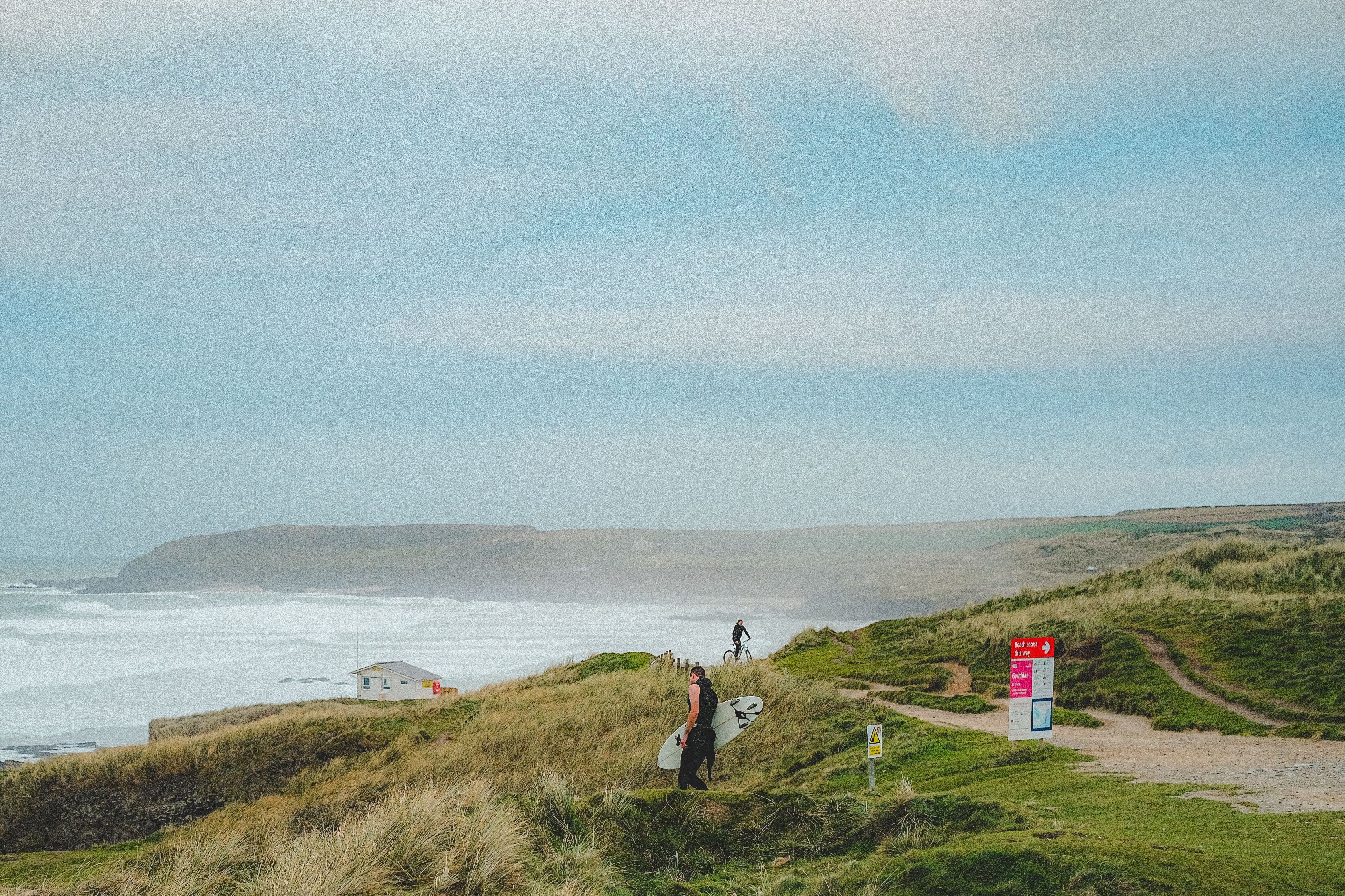 Surfer carrying a board along the grassy dunes above a Cornish beach