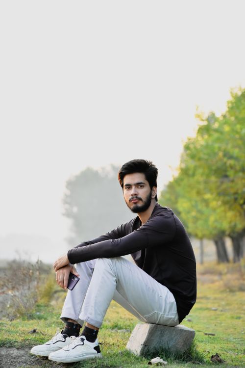 Free Young man with a beard sitting on a rock outdoors, holding a smartphone, surrounded by greenery. Stock Photo