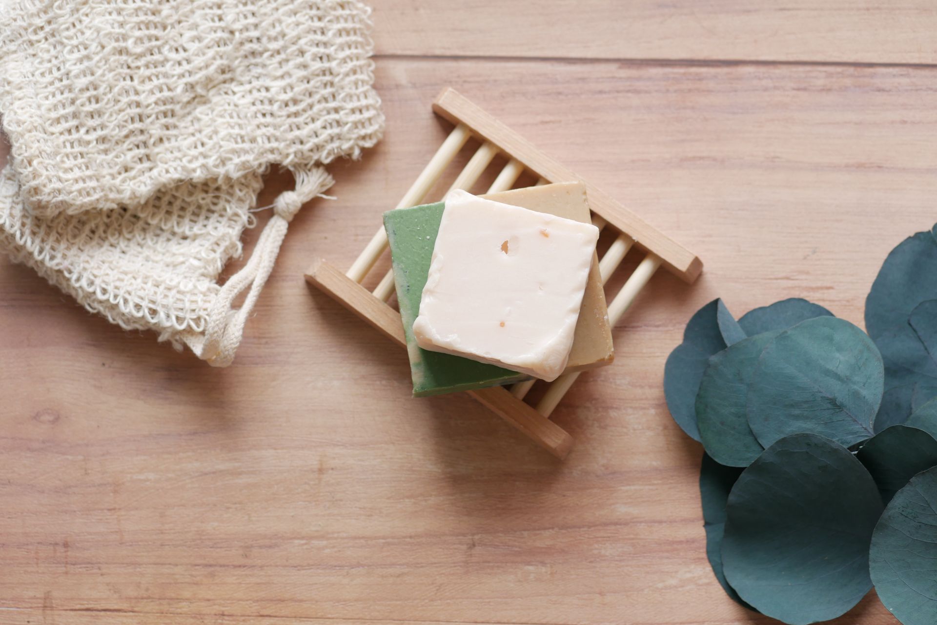 Top view of handcrafted soap bars on bamboo dish with green leaves, featuring a rustic wooden background.