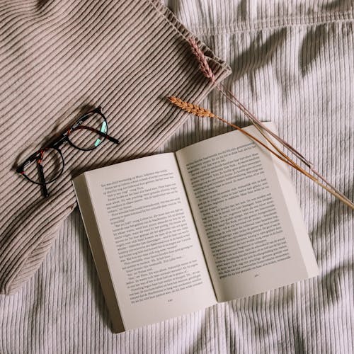 An Open Book Beside Eyeglasses on Brown Fabric