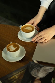 Hands hold coffee cups with latte art on a wooden table beside a laptop.