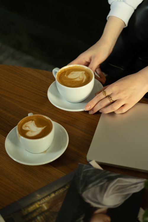 Free Hands hold coffee cups with latte art on a wooden table beside a laptop. Stock Photo
