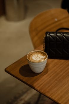 A cozy scene with a latte in a ceramic cup on a wooden table beside a black leather bag.