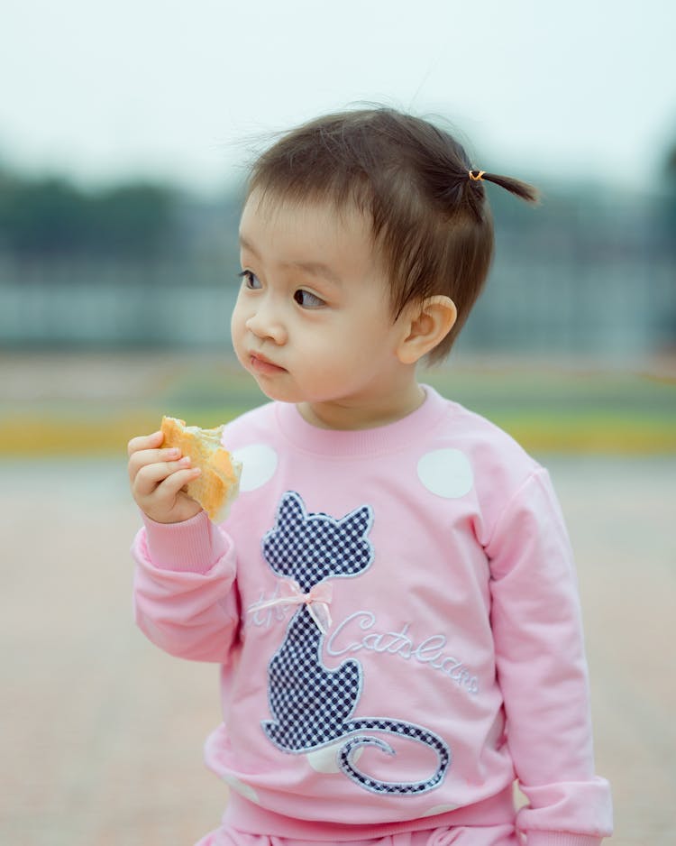Baby Girl Wearing Pink Blouse Eating A Bread