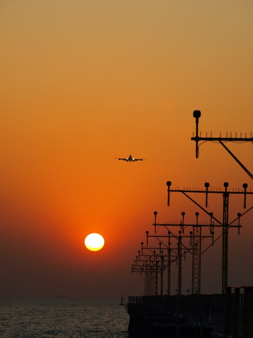 A Silhouette of an Airplane Flying over the Electricity Post Near Body of Water During Sunset