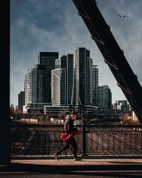 A man walks across a bridge with a skyline of modern high-rise buildings under a clear sky.