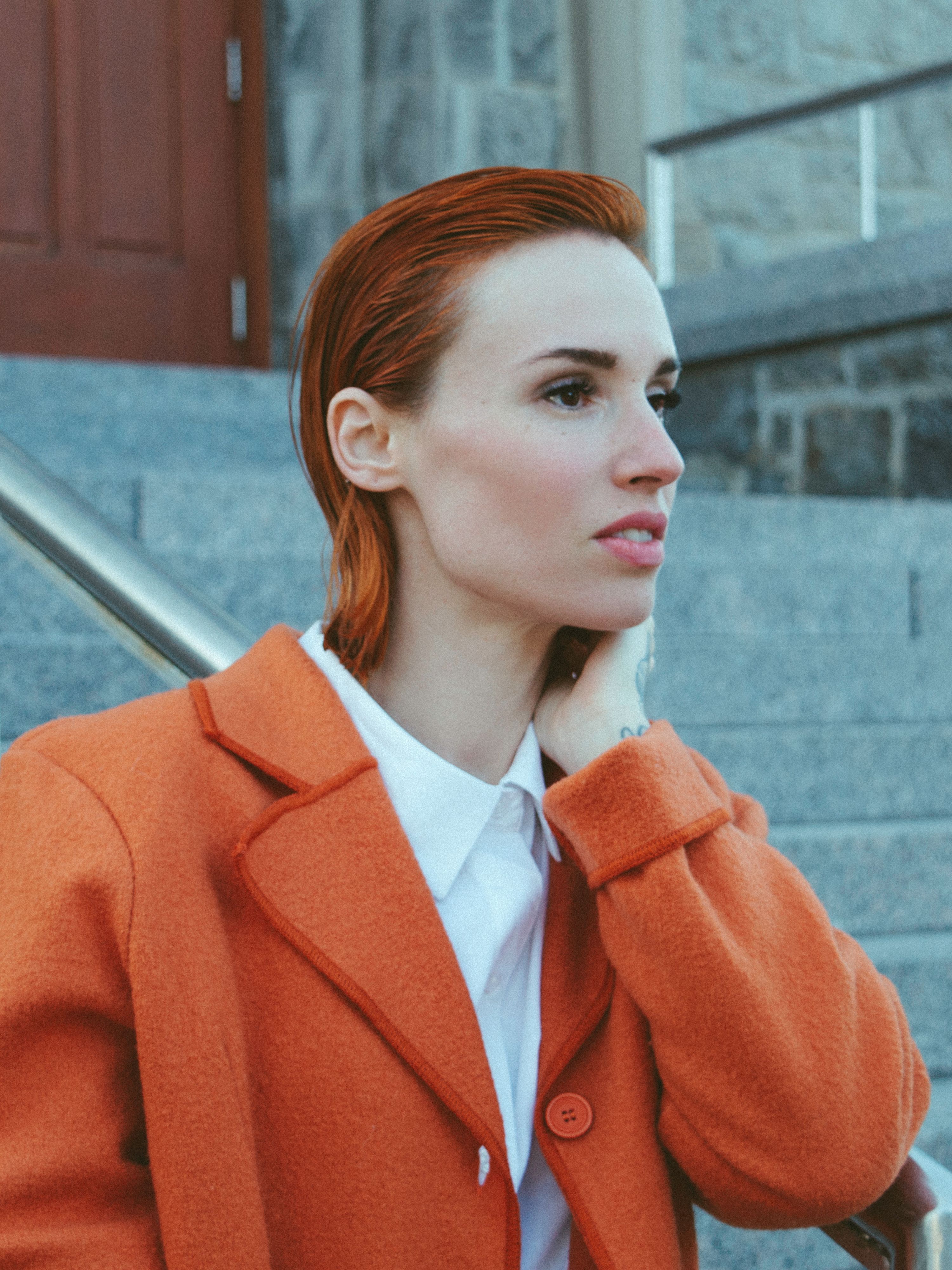 Portrait of a woman with red hair in an orange coat against a stone building backdrop.