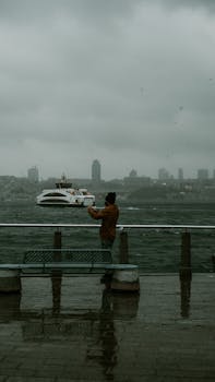 A solitary man stands by the seaside on a foggy day, watching a ferry pass.