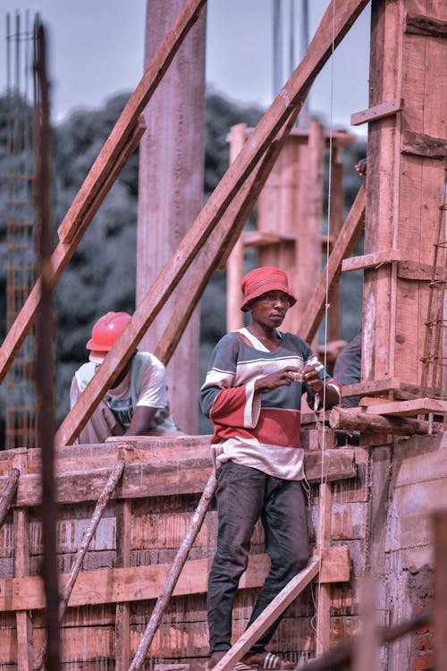 A Man in Red and Blue Shirt Leaning a Wooden Structure Near a Person with Red Helmet