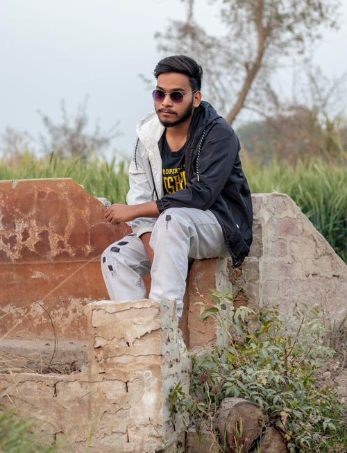 Free A young man in sunglasses sitting on a concrete wall in a natural outdoor setting. Stock Photo
