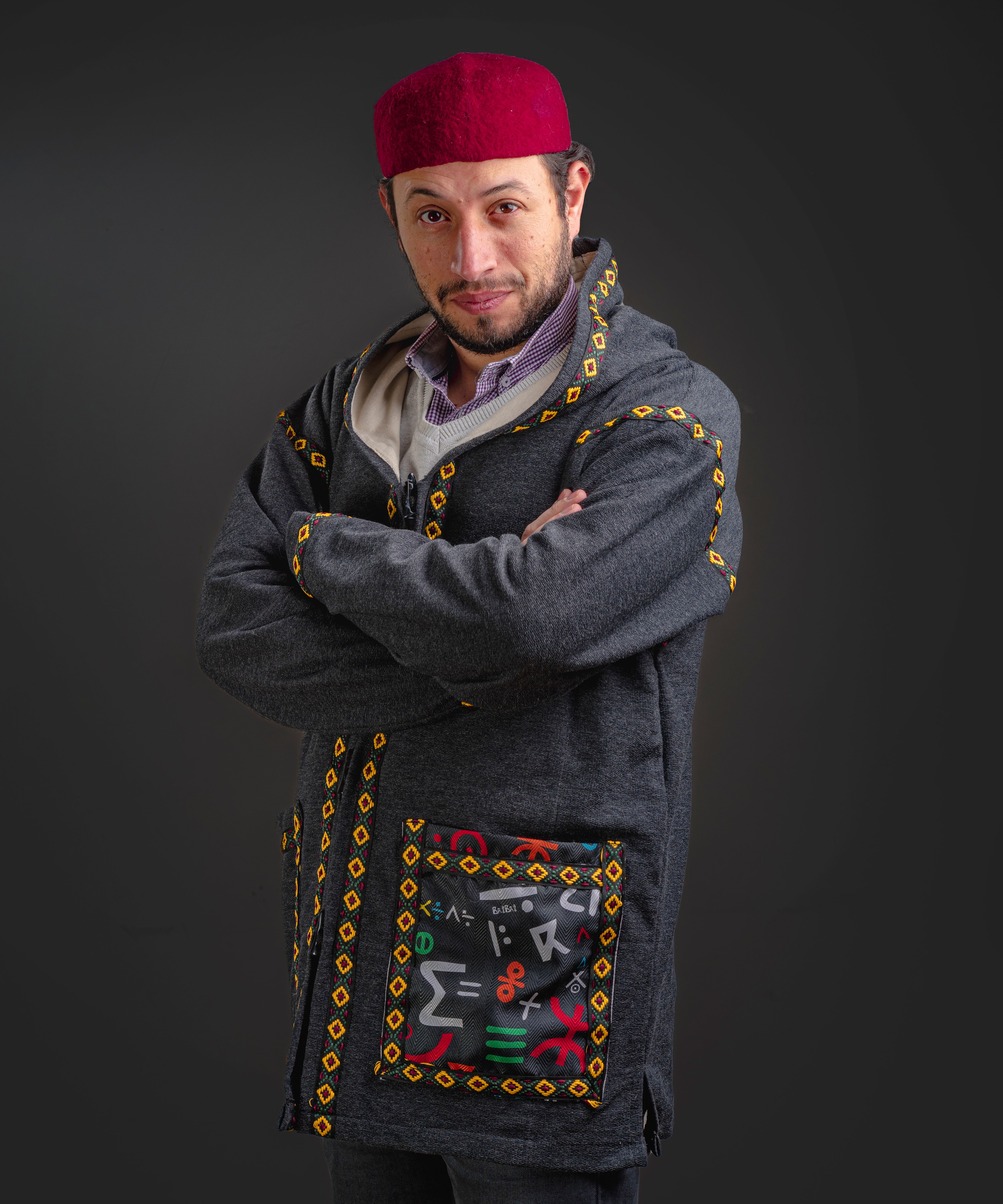 Man wearing a red hat and traditional embroidery adorned coat, arms crossed, studio portrait.