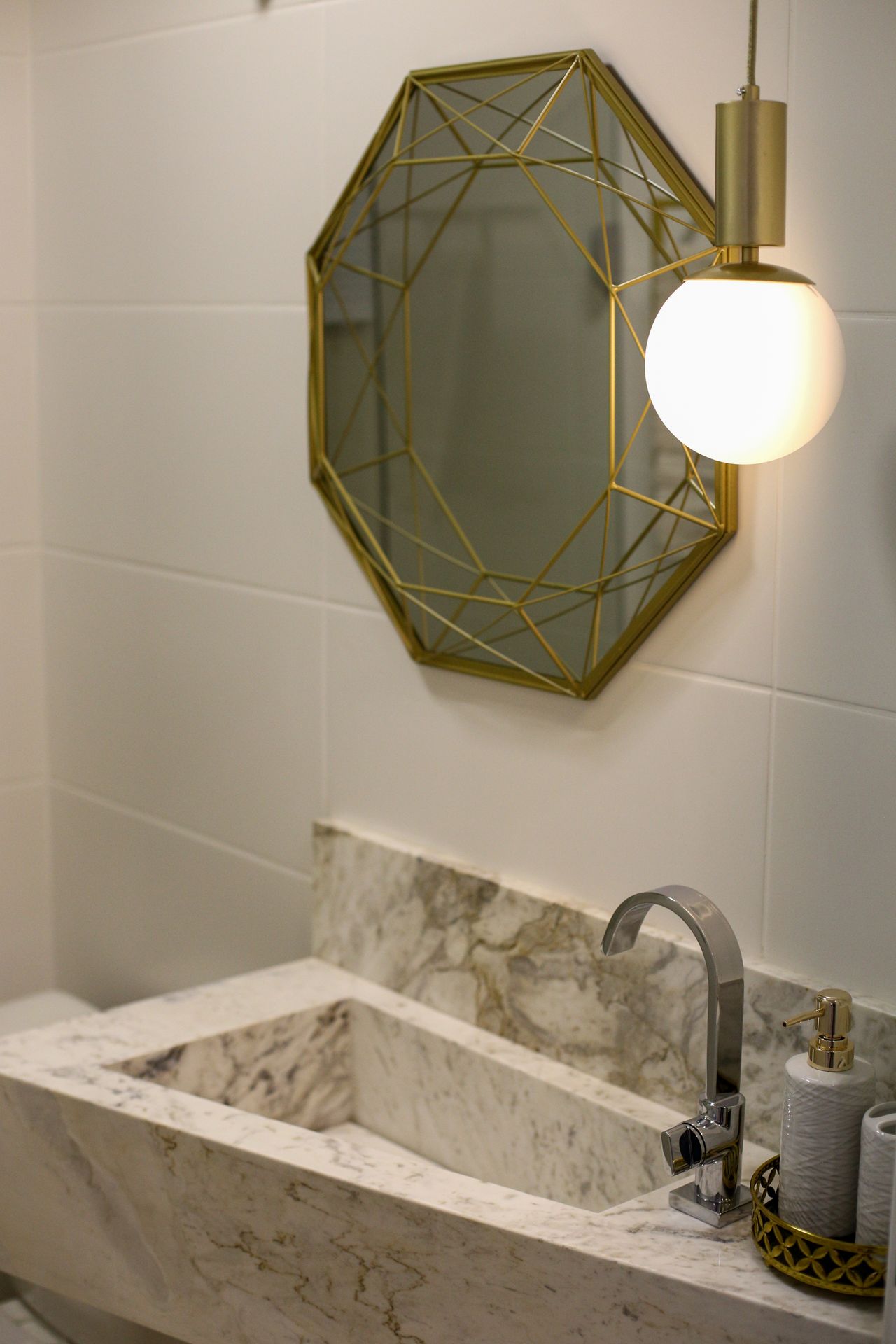 Brass pendant light with a white globe hanging beside a geometric gold mirror over a marble bathroom sink