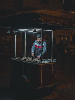 A street vendor sells roasted chestnuts under illuminated cart, city night scene.