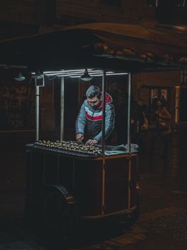 A street vendor prepares chestnuts under bright lights during a bustling night market.