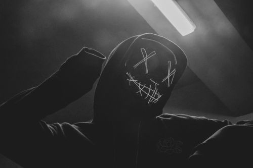 Free A mysterious person wearing a neon mask and hoodie in a shadowy environment. Stock Photo