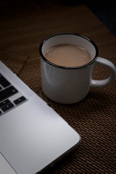 Rustic white coffee mug with latte next to a laptop on a textured woven mat, ideal for cozy workspaces.