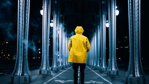 Free Person in a yellow raincoat stands alone on a bridge at night, surrounded by industrial architecture. Stock Photo