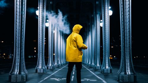 Free A person in a yellow jacket stands on a night-lit bridge walkway under arches and columns. Stock Photo