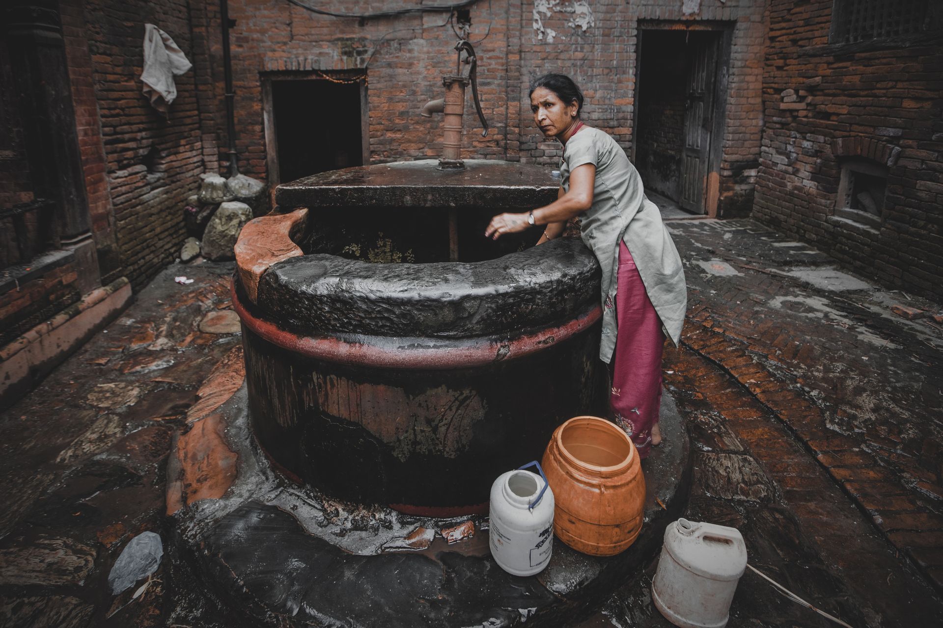 A woman in a traditional courtyard gathering water from a rustic well, surrounded by containers.