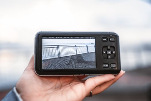 Close-up of a hand holding a digital camera showing a view of a cityscape railing.