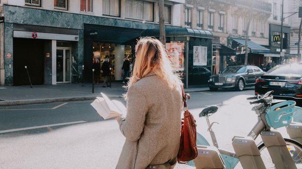 A woman reads a book while standing on a lively city street with shops and parked cars.