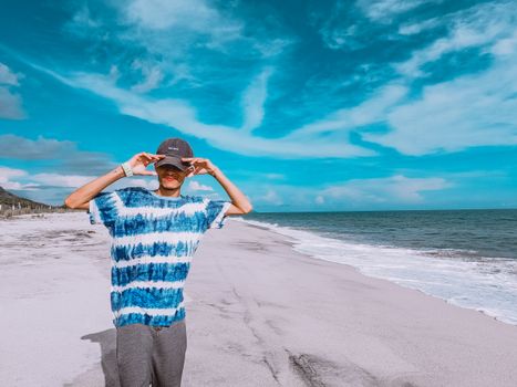A man in a tie-dye shirt poses on a serene beach with a vibrant blue sky.