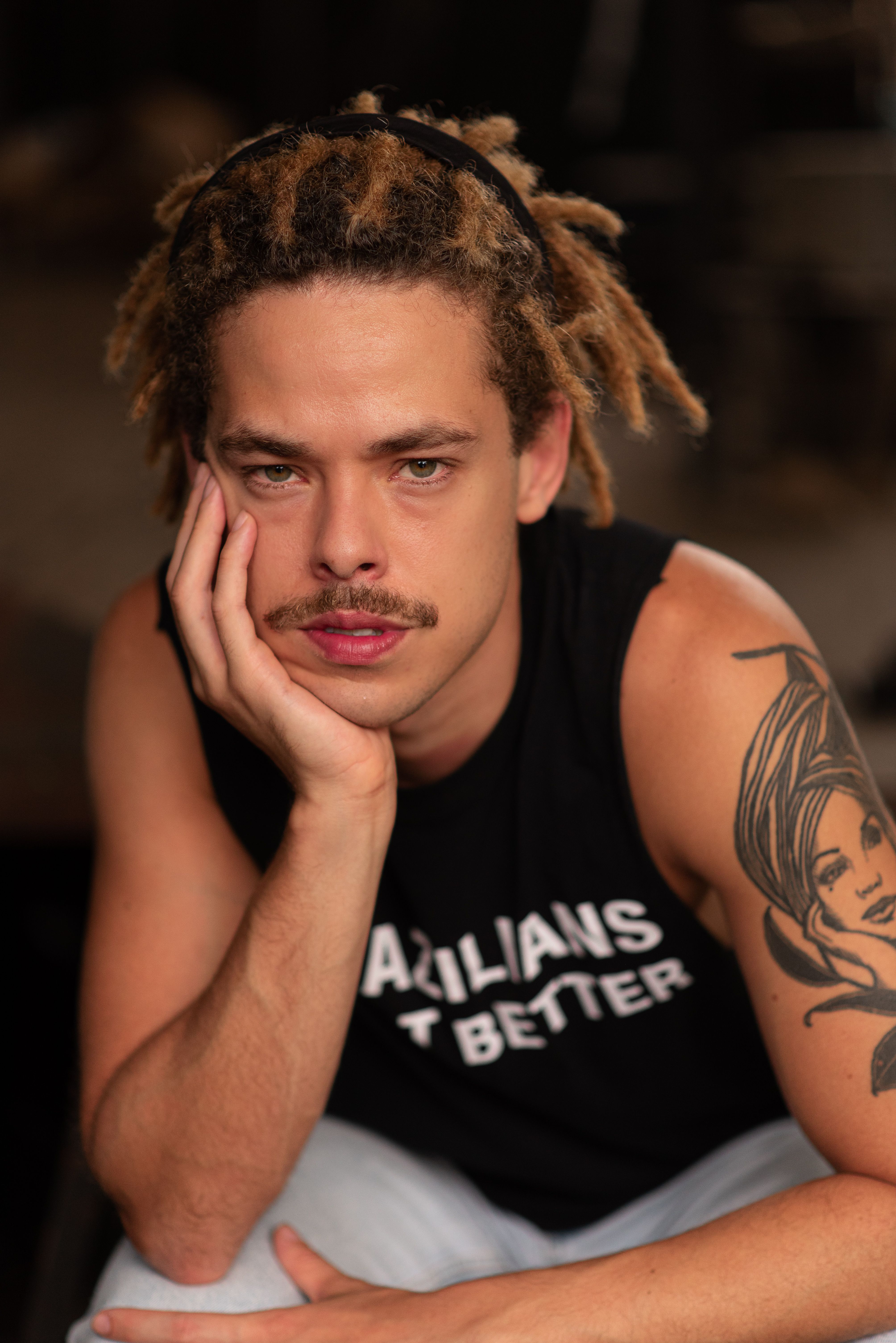 Close-up portrait of a man with dreadlocks and arm tattoo posing thoughtfully.