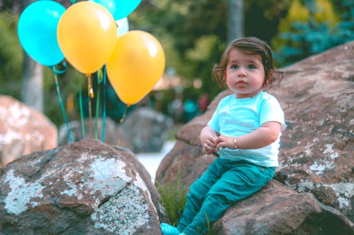 Toddler Wearing White Shirt Sitting on Rock Beside Yellow and Blue Balloons