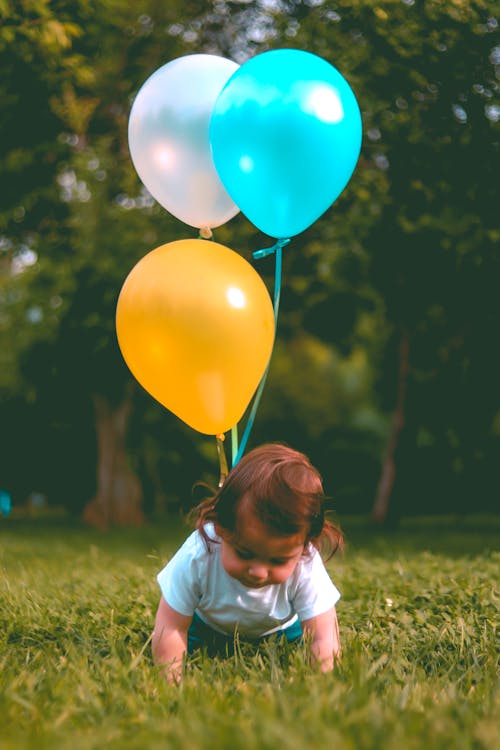 嬰兒穿白色t卹，在樹林附近的綠色草地上抱著三個黃色，藍色和白色的氣球