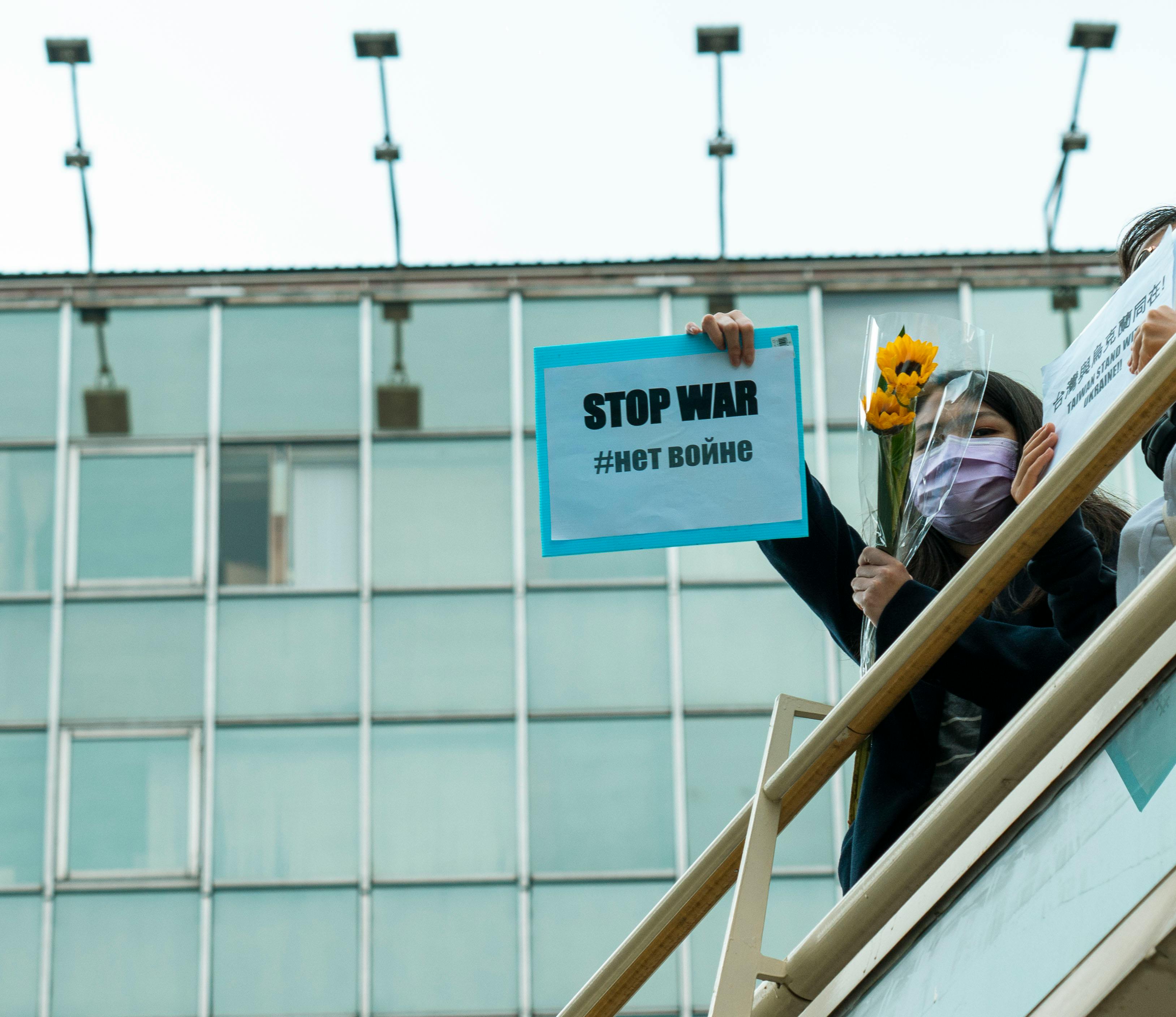 A woman with a mask holds a 'Stop War' sign and a sunflower at an outdoor protest.