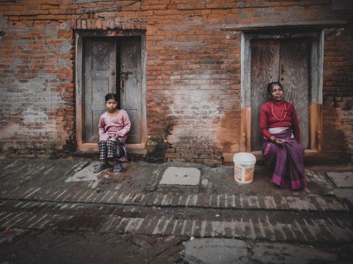 Free Two women sitting on doorsteps against a brick wall in an urban setting, exuding an earthy and moody atmosphere. Stock Photo