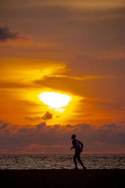 Unternehmer Training am Bodensee A man runs along the beach in Angola, silhouetted against a vibrant sunset sky over the tranquil ocean.