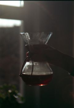 A silhouette image of a hand holding a Chemex coffee maker with light streaming through a window.