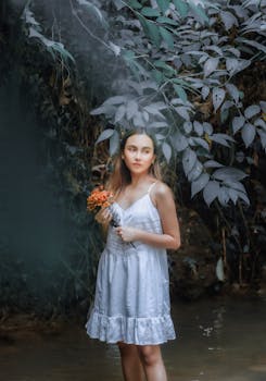 A young woman in a white dress holds a bouquet of orange flowers in a tranquil forest setting.