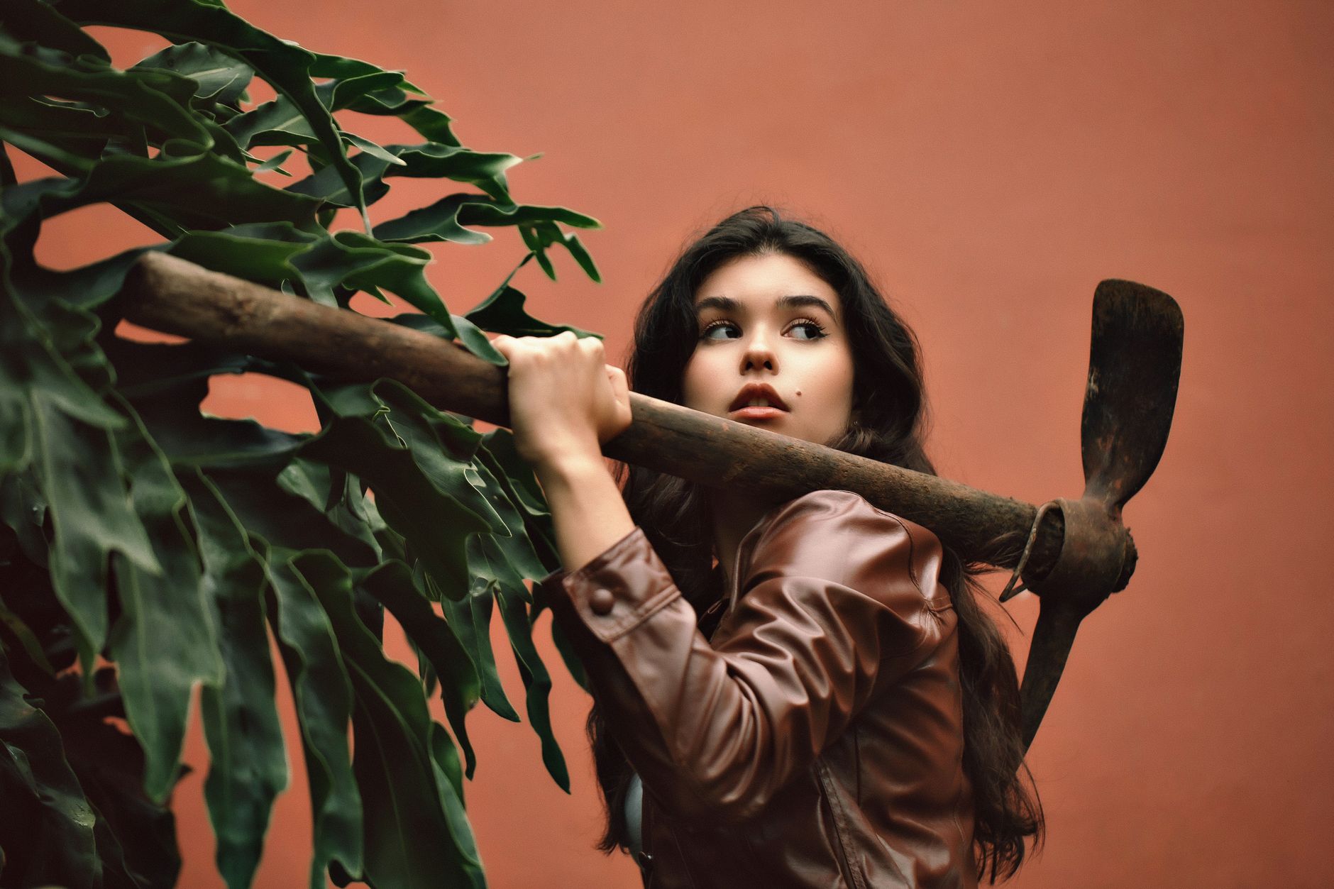 Portrait of a woman posing with a mattock, wearing a brown leather jacket against a terracotta background.