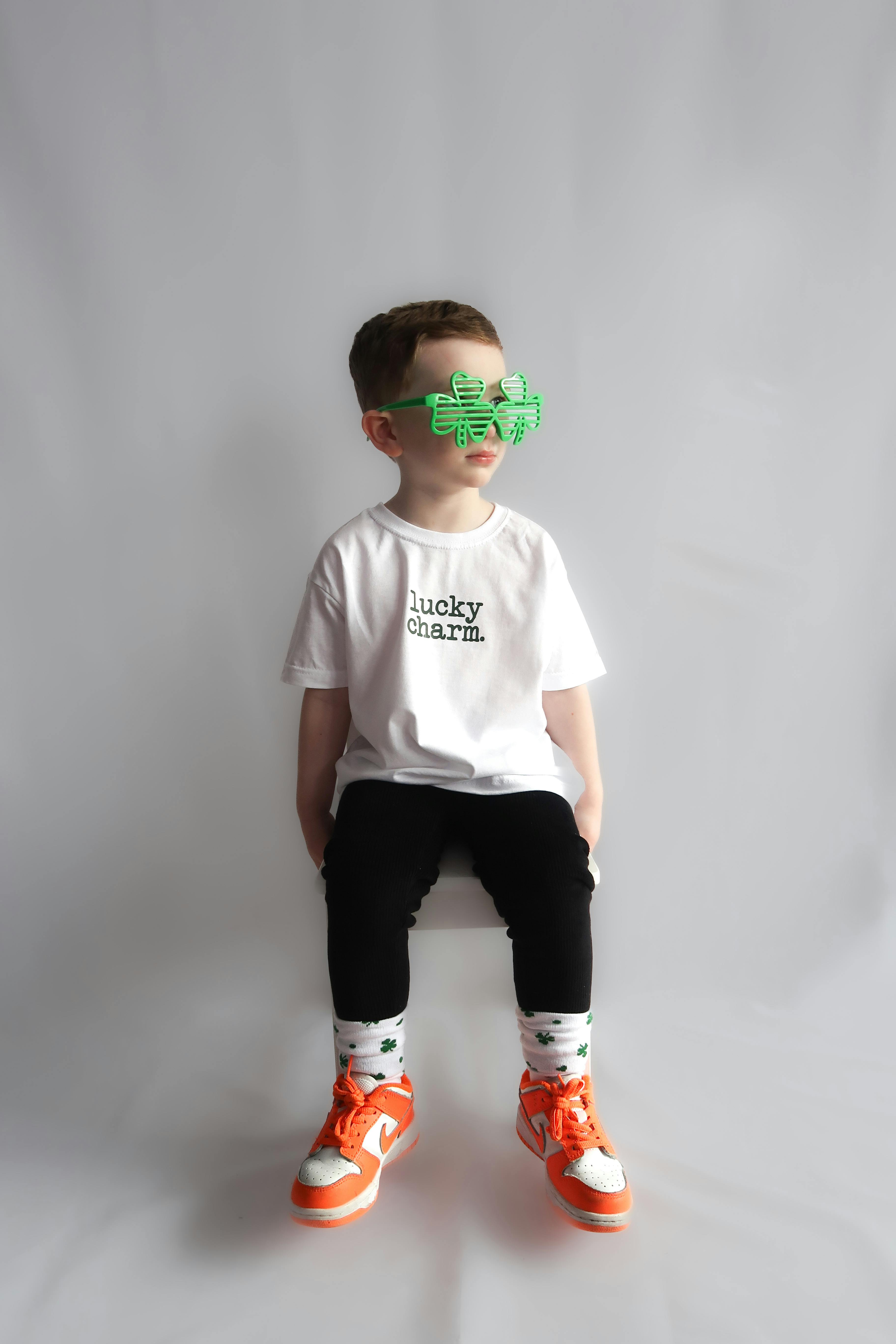 Adorable child in festive St. Patrick's Day outfit with shamrock glasses, sitting against a plain background.