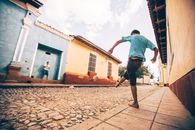 A young boy kicks a soccer ball in a colorful cobblestone street on a sunny day.