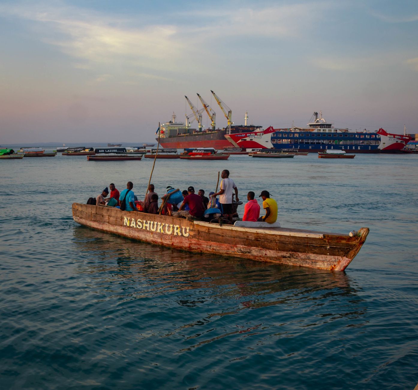 A group of people on a traditional wooden boat in Zanzibar at sunset, with ships in the background.