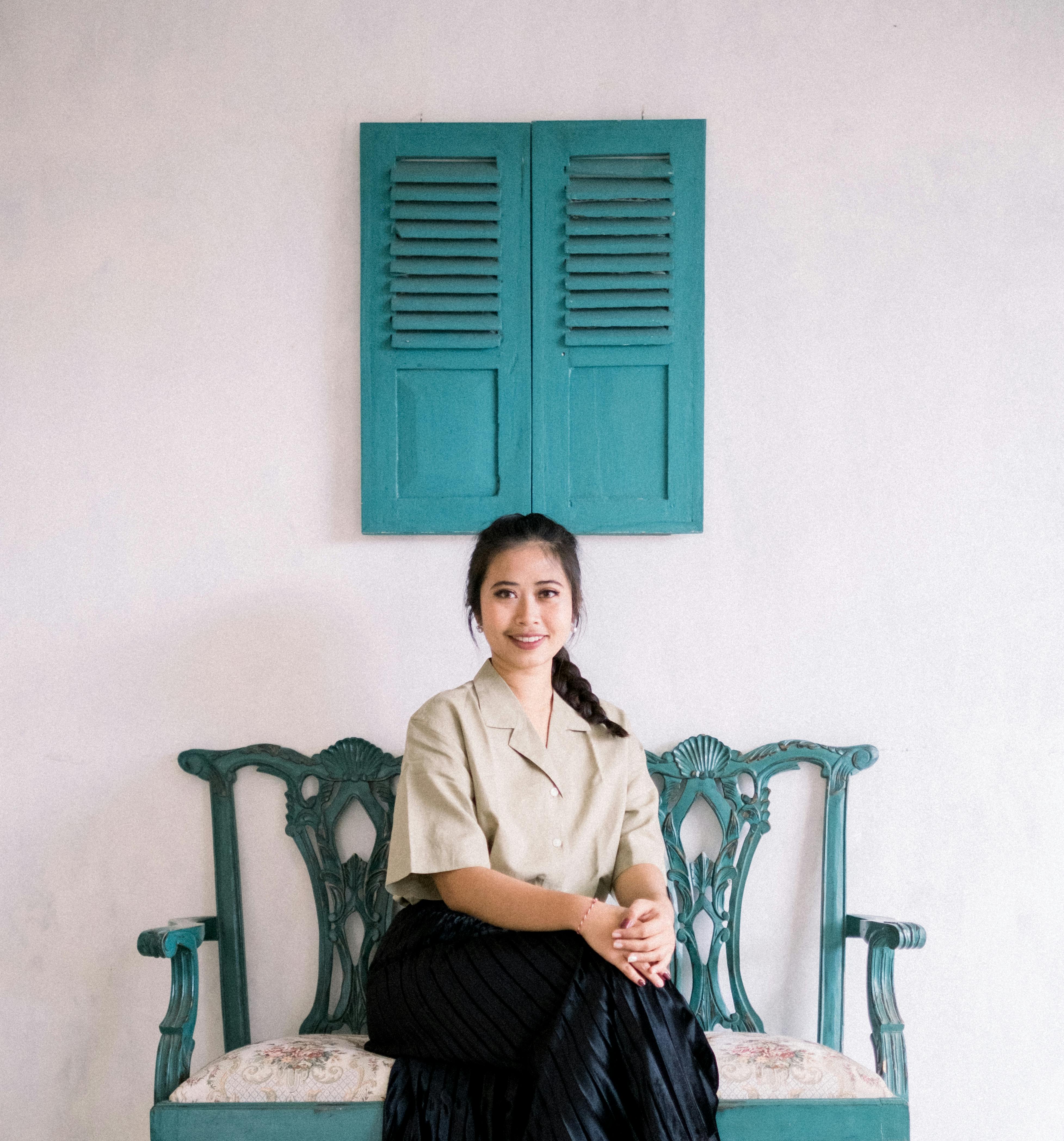 A woman in a black skirt sits elegantly on a vintage bench, smiling warmly indoors.