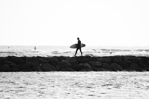 Grayscale Photo of Man Walking on Jetty While Holding Surfboard