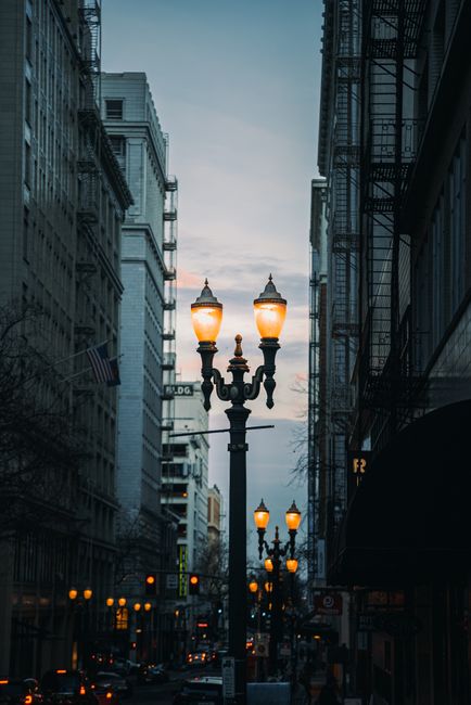 A city street flanked by tall buildings and glowing vintage lamp posts at twilight.