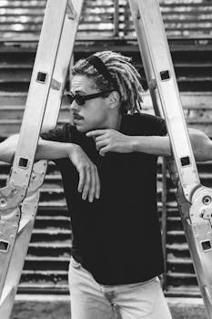 Black and white portrait of a stylish man with dreadlocks leaning on a ladder.
