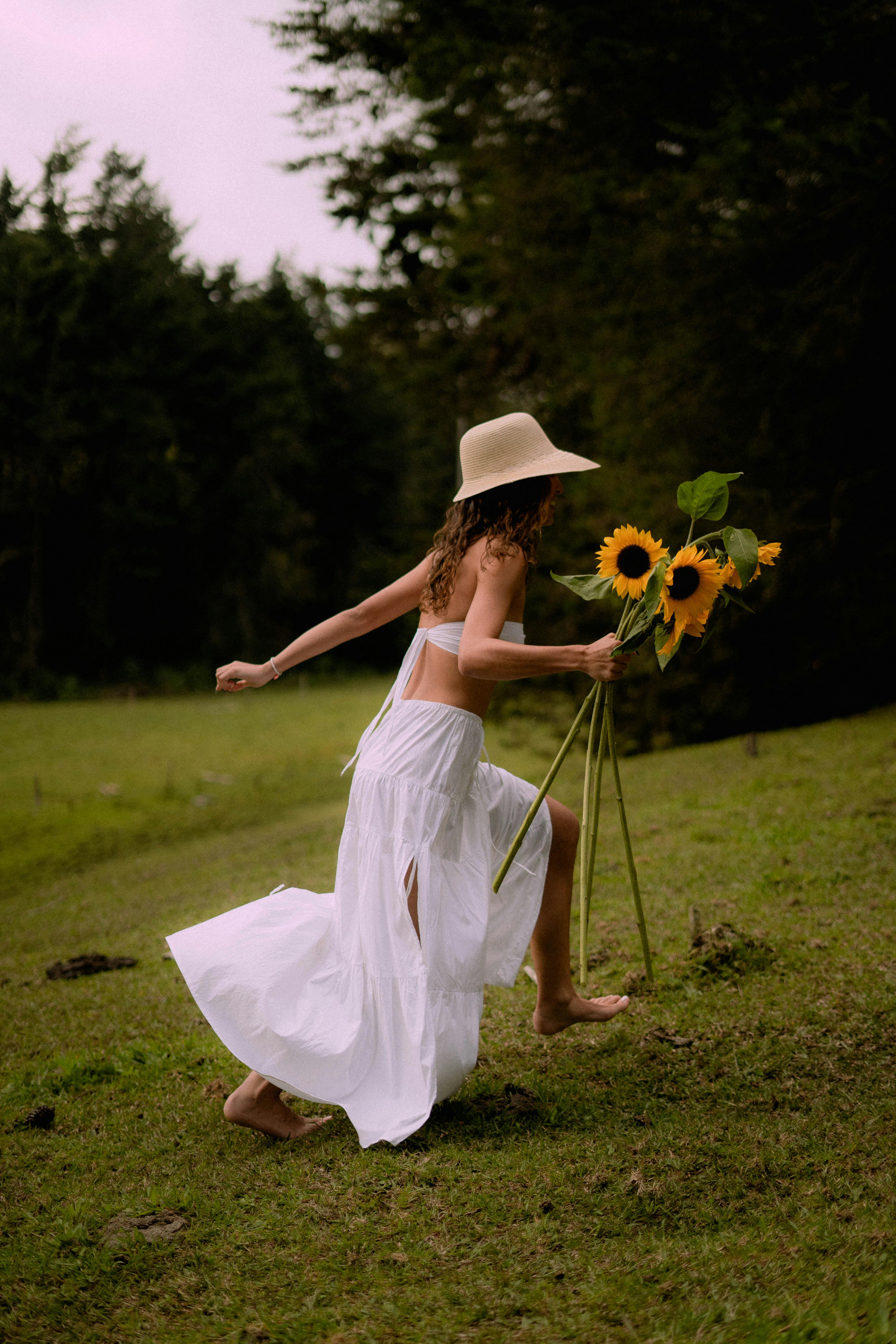 A woman in a white skirt carrying sunflowers runs barefooted on a green field.
