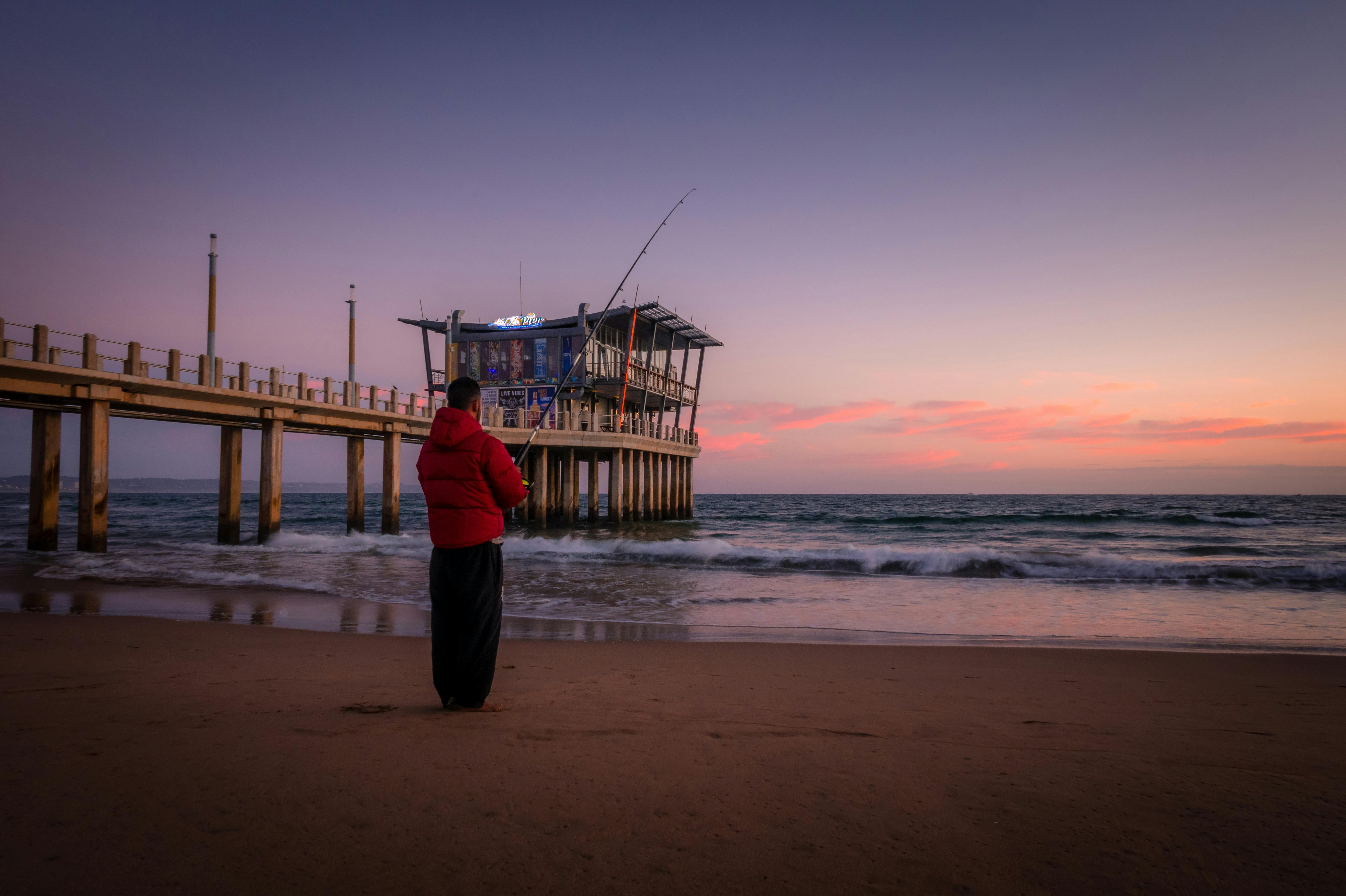 Man in red jacket fishing at Durban seashore with scenic twilight sky and waves.