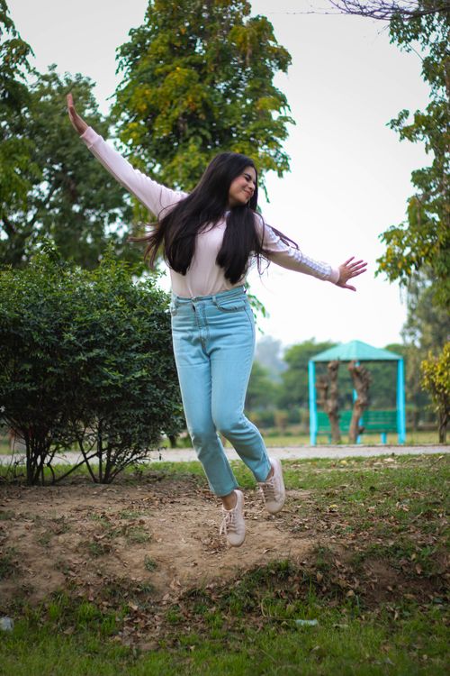 Free Young woman with long hair jumping joyfully in a park, wearing jeans and a long sleeve shirt. Stock Photo