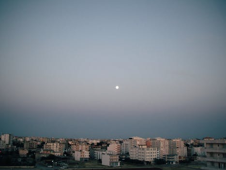 A serene aerial view of a cityscape under the evening sky with the moon visible.