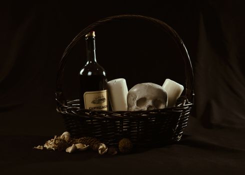 A moody still life featuring a wine bottle, skull, and candles in a basket.