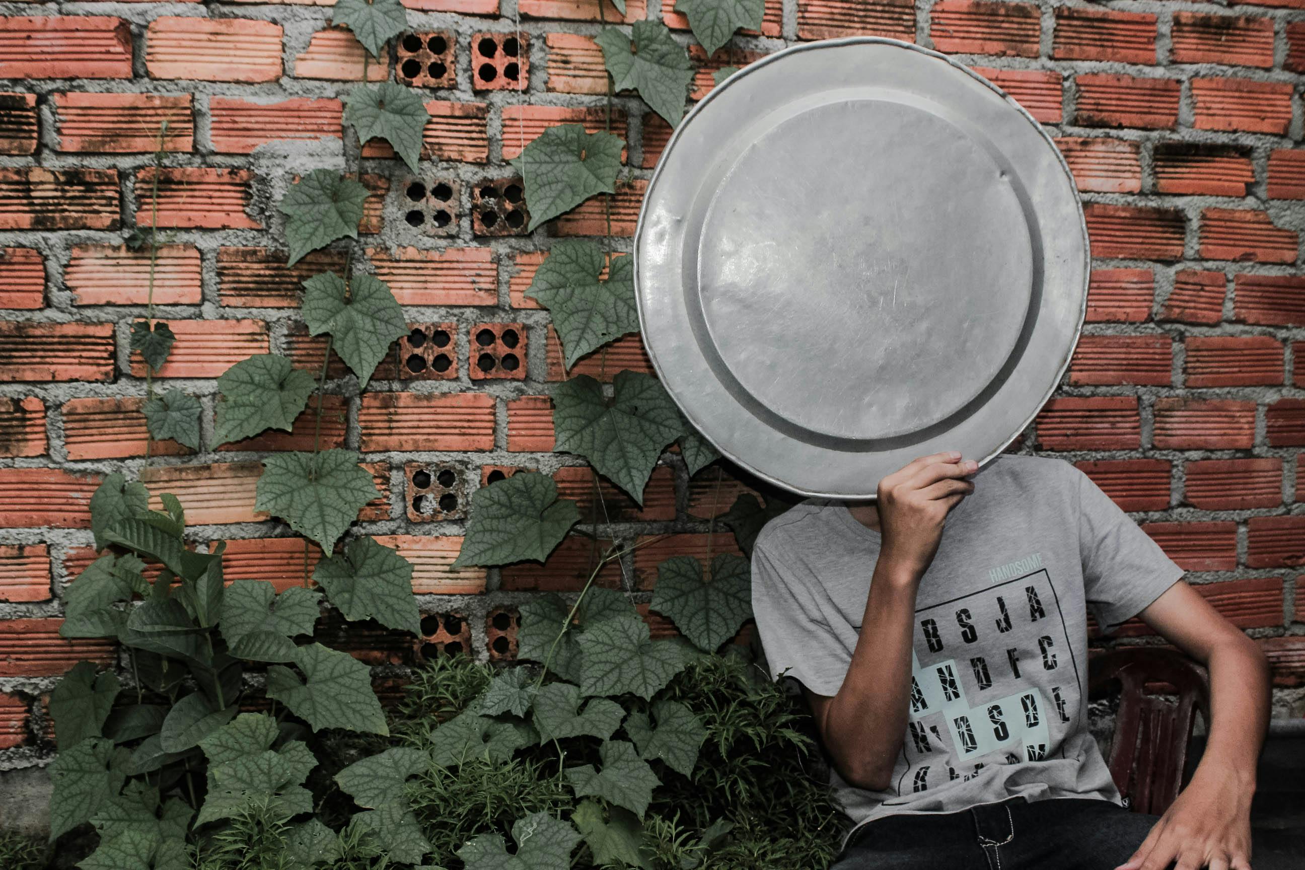 一个人用金属托盘遮住脸，站在覆盖着绿色藤蔓的砖墙前