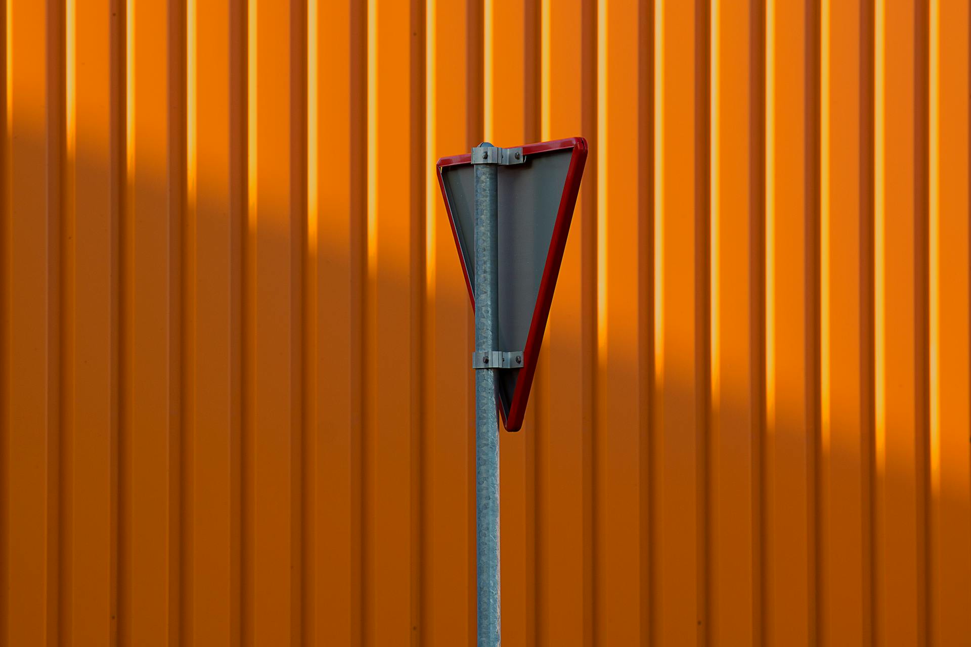 Back view of a triangular traffic sign against a vibrant orange corrugated wall.